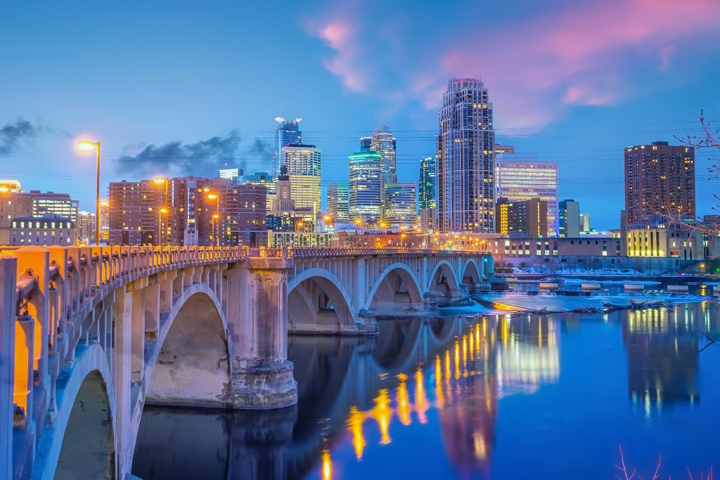 City skyline at dusk with an arched bridge over a reflective river and glowing streetlights.