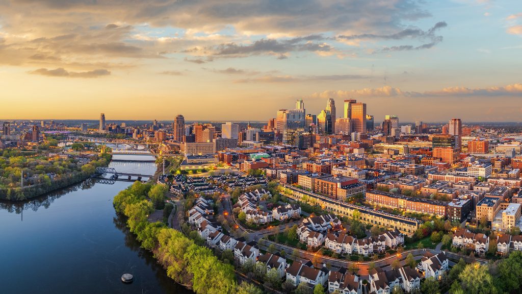 Aerial view of a city skyline at sunset with a river, bridges, and a mix of tall buildings and suburban neighborhoods in the foreground.