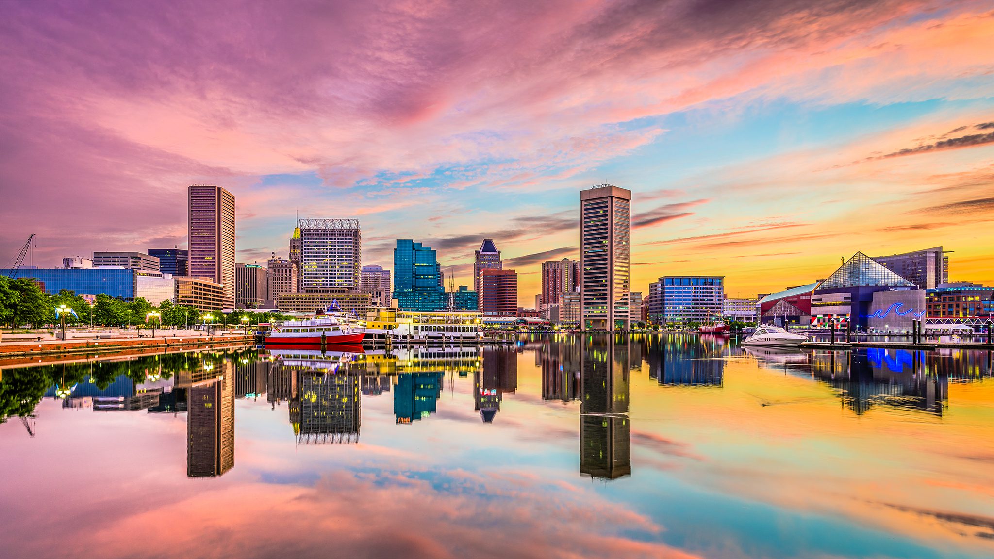 City skyline along a harbor at sunset with moored boats and a vibrant pink-to-orange sky reflecting on calm water.
