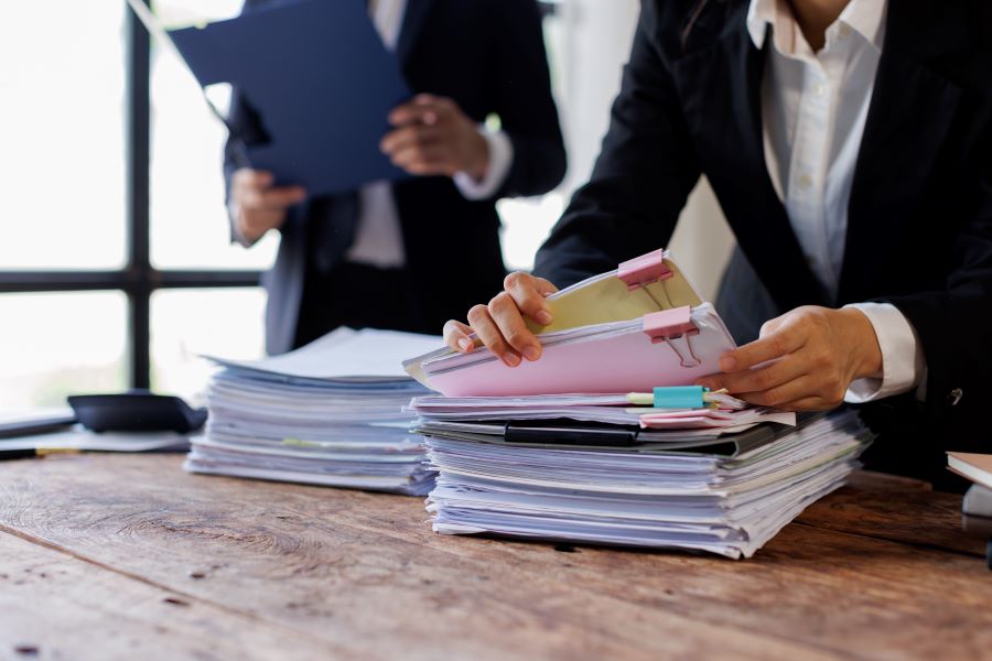 Two professionals in business attire sort through a tall stack of documents on a wooden desk, clipping pink folders