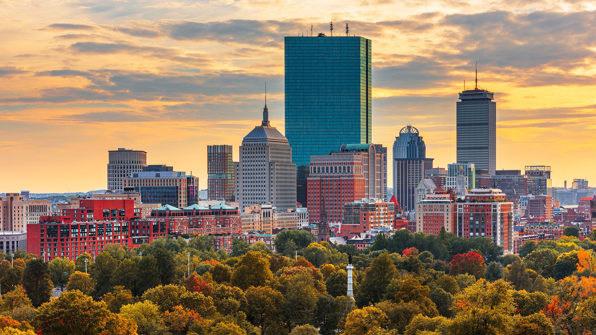 City skyline at sunset with a tall blue glass skyscraper and surrounding buildings, framed by autumn-colored trees in the foreground.