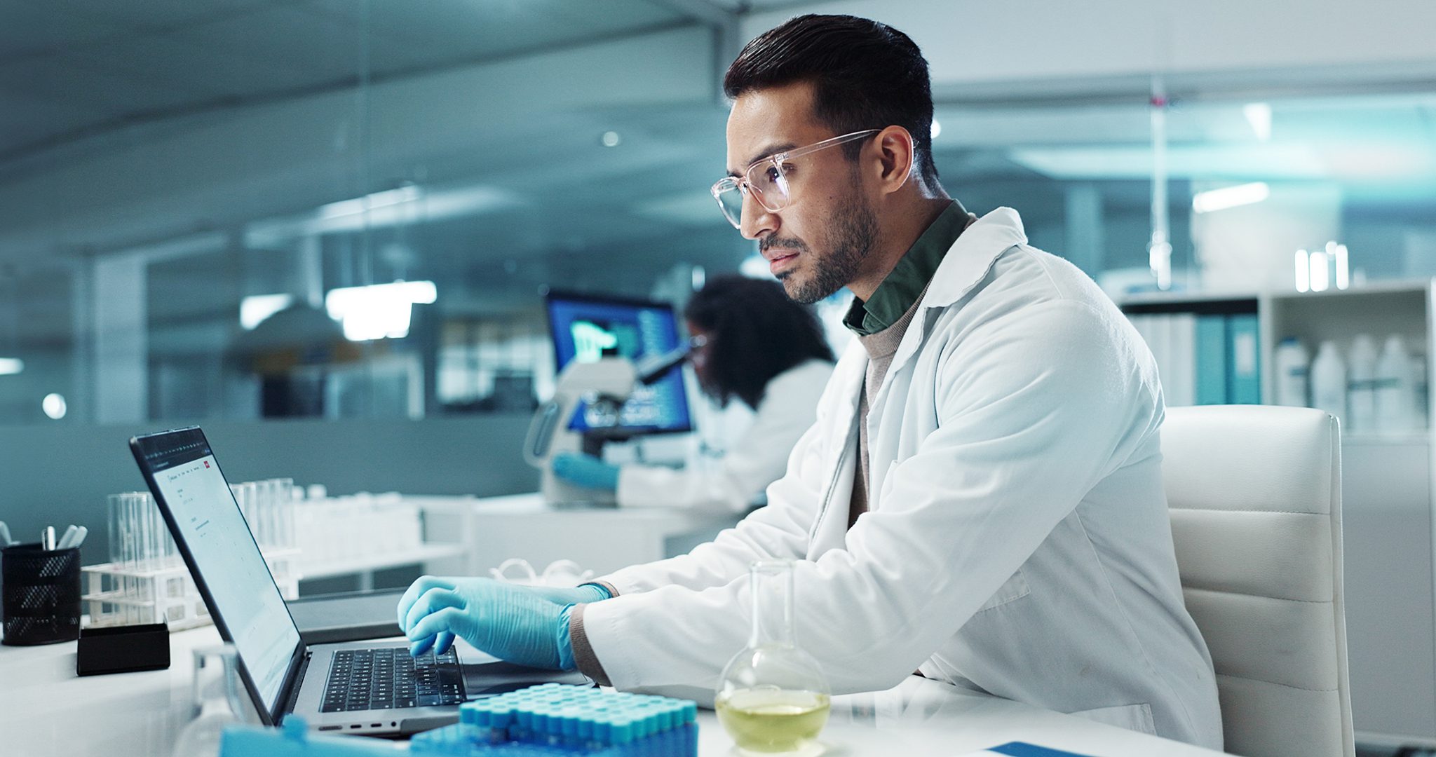 Scientist in a white lab coat and safety glasses types on a laptop at a lab bench with test tubes nearby.