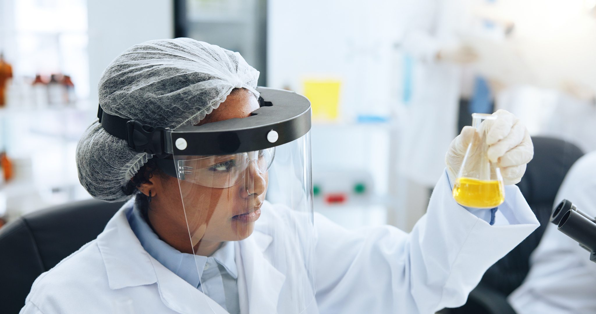 Female scientist in a lab coat, hairnet, and face shield holds a yellow liquid in a beaker.